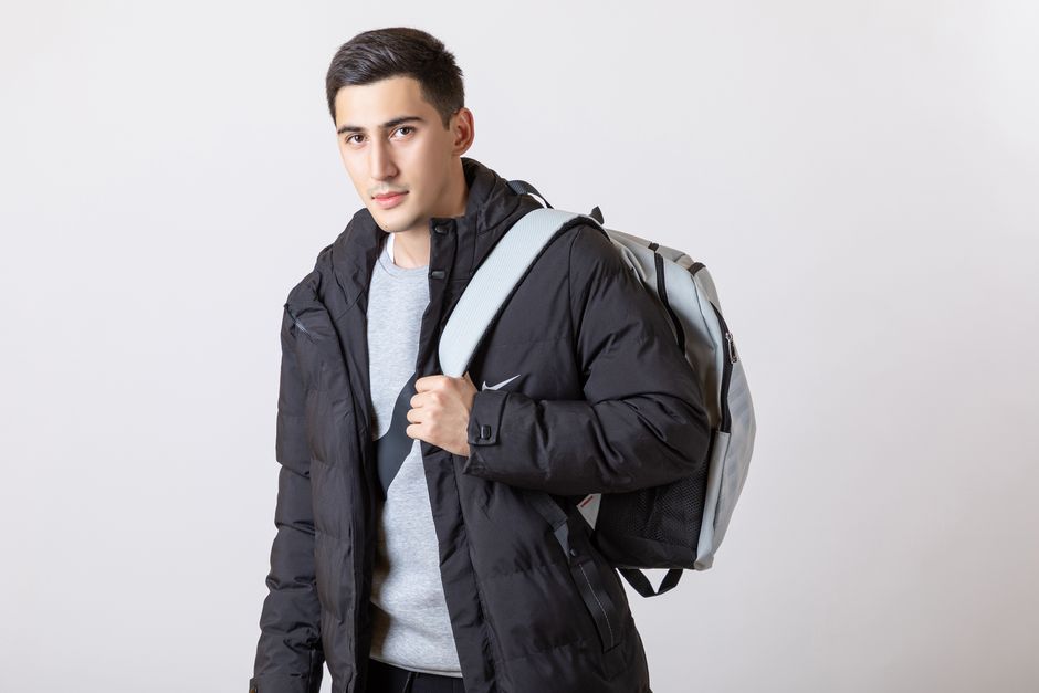 A stylish man posing in a studio wearing casual outdoor fashion with a backpack.