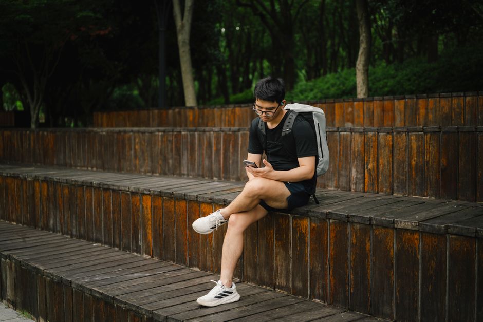 A young man sits on wooden benches outdoors, using a smartphone and wearing a casual outfit with a backpack.